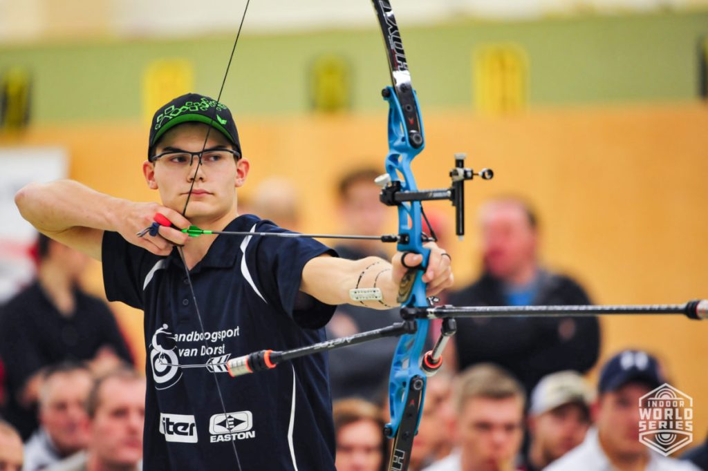 Een boogschutter met een bril mikt op een boog tijdens een indoor handboogsportwedstrijd. Op de achtergrond zijn toeschouwers zichtbaar.