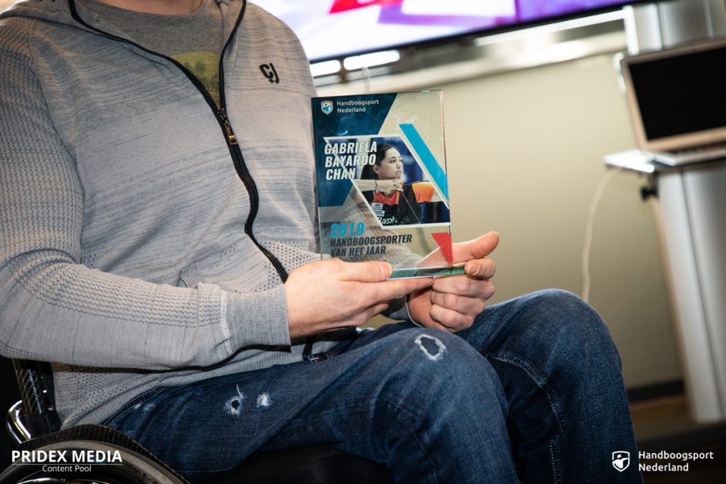 Een persoon in een rolstoel houdt een bord vast met de titel "Gabriela Bayardo Chan 2019 Handboogsporter van het Jaar" voor een PRIDEX MEDIA-banner met een laptop en pijl op de achtergrond.