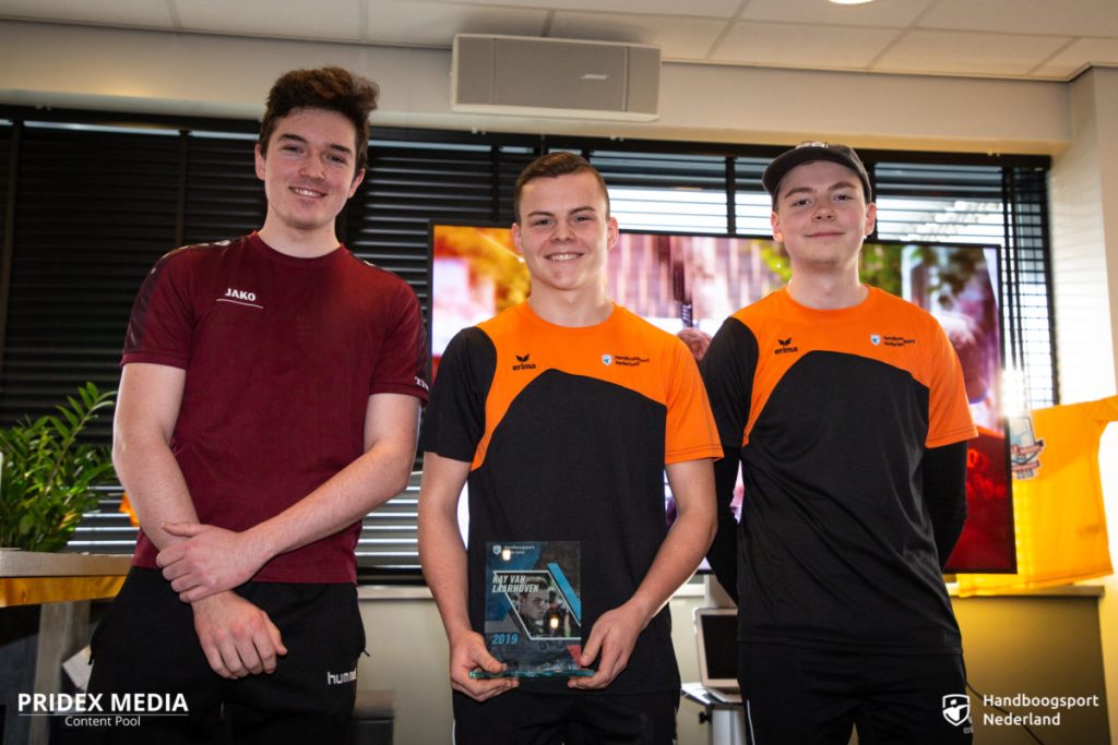 Drie jonge mannen staan binnen en glimlachen naar de camera. Eén houdt een glazen trofee vast, waarmee hun triomf in de handboogsport wordt getoond. Ze dragen allemaal sportuniformen, waarvan twee in bijpassende zwarte en oranje shirts. Logo's op de achtergrond duiden op een sportevenement.