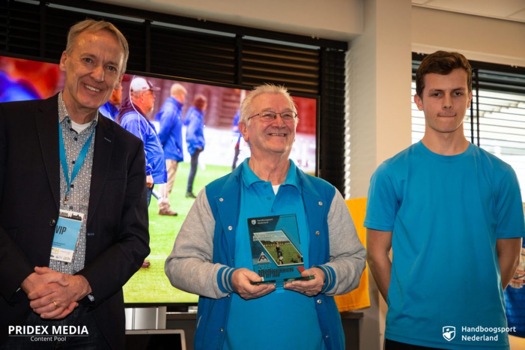 Drie mannen staan binnen; degene in het midden houdt een plaquette vast. Links staat een man in pak glimlachend, en rechts staat een jongeman in een blauw overhemd neutraal. Op de achtergrond staat een scherm met mensen erop, waarop wellicht hoogtepunten van recente handboogsportevenementen te zien zijn.