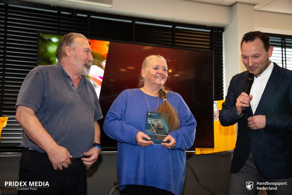 Drie mensen staan voor een scherm, waarbij de middelste een plaquette vasthoudt. De persoon rechts spreekt in een microfoon en bespreekt handboogsport. Linksonder staat het logo "PRIDEX MEDIA" en rechtsonder staat "Handboogsport Nederland".