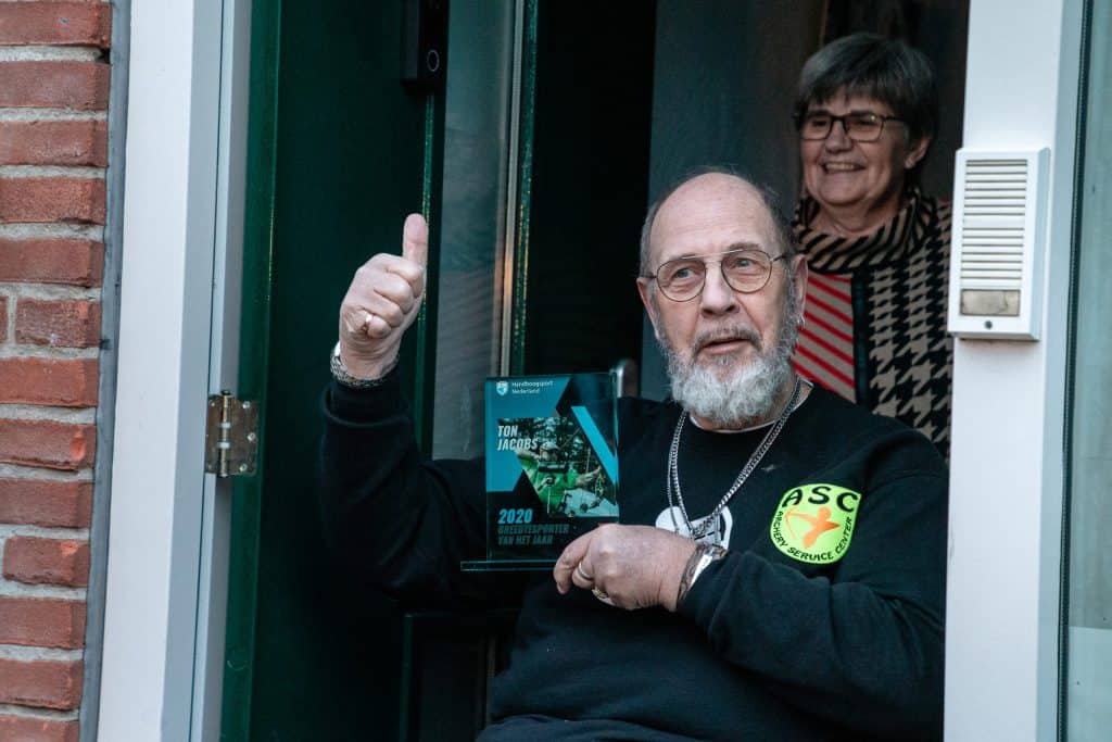 Een oudere man met een bril en een baard zit in een deuropening, houdt een plaquette vast en steekt zijn duim omhoog. Achter hem staat een vrouw, trots glimlachend. De plaquette herdenkt zijn indrukwekkende handboogschietvaardigheid.