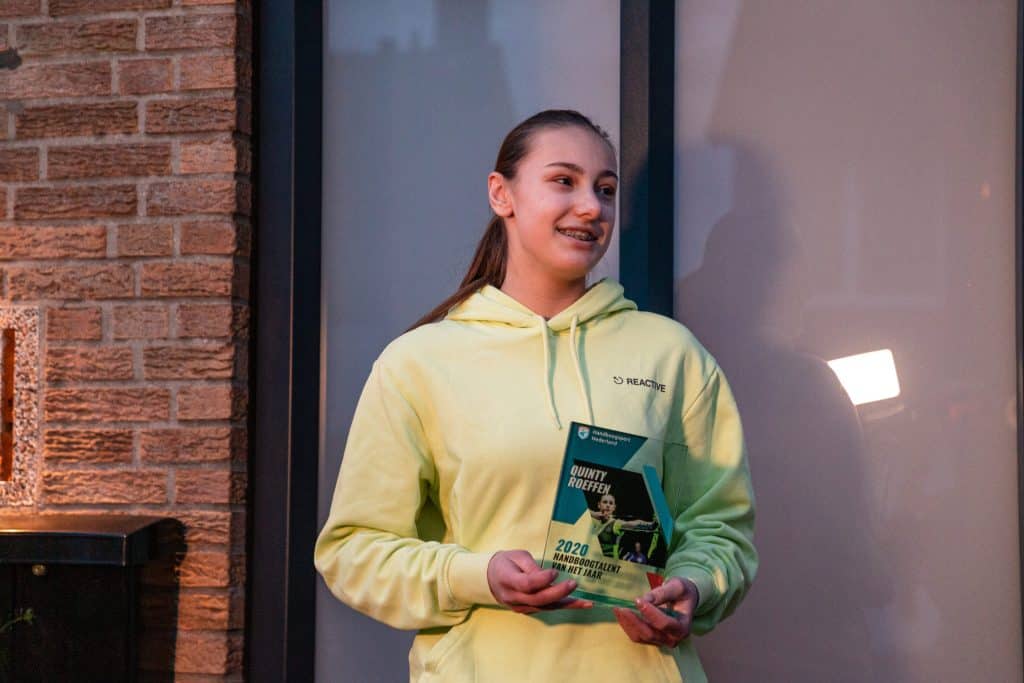 Een persoon in een gele hoodie glimlacht terwijl hij een trofee voor een bakstenen muur en glazen deur houdt en viert trots zijn overwinning met het handboogschieten.