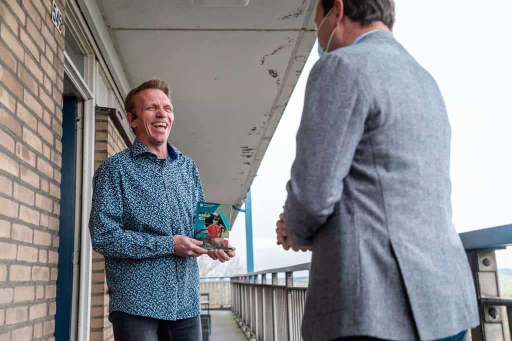 Een man op een balkon glimlacht en houdt een product vast, terwijl hij de fijne kneepjes van het handboogschieten bespreekt met iemand anders die met zijn rug naar de camera staat.