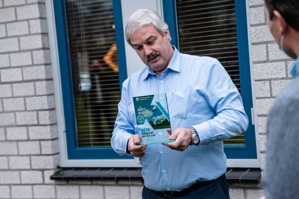 Een man met grijs haar en een snor houdt een plaquette met beide handen vast. Hij staat voor een raam naast een bakstenen muur. Rechts is gedeeltelijk nog een persoon zichtbaar, mogelijk betrokken bij handboogsport.