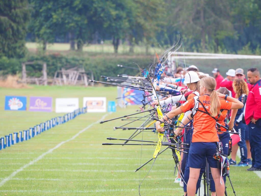 Boogschutters in oranje shirts staan in een rij, richten hun bogen en richten hun pijl op doelen in een buitenveld met bomen op de achtergrond, terwijl ze handboogschieten oefenen.