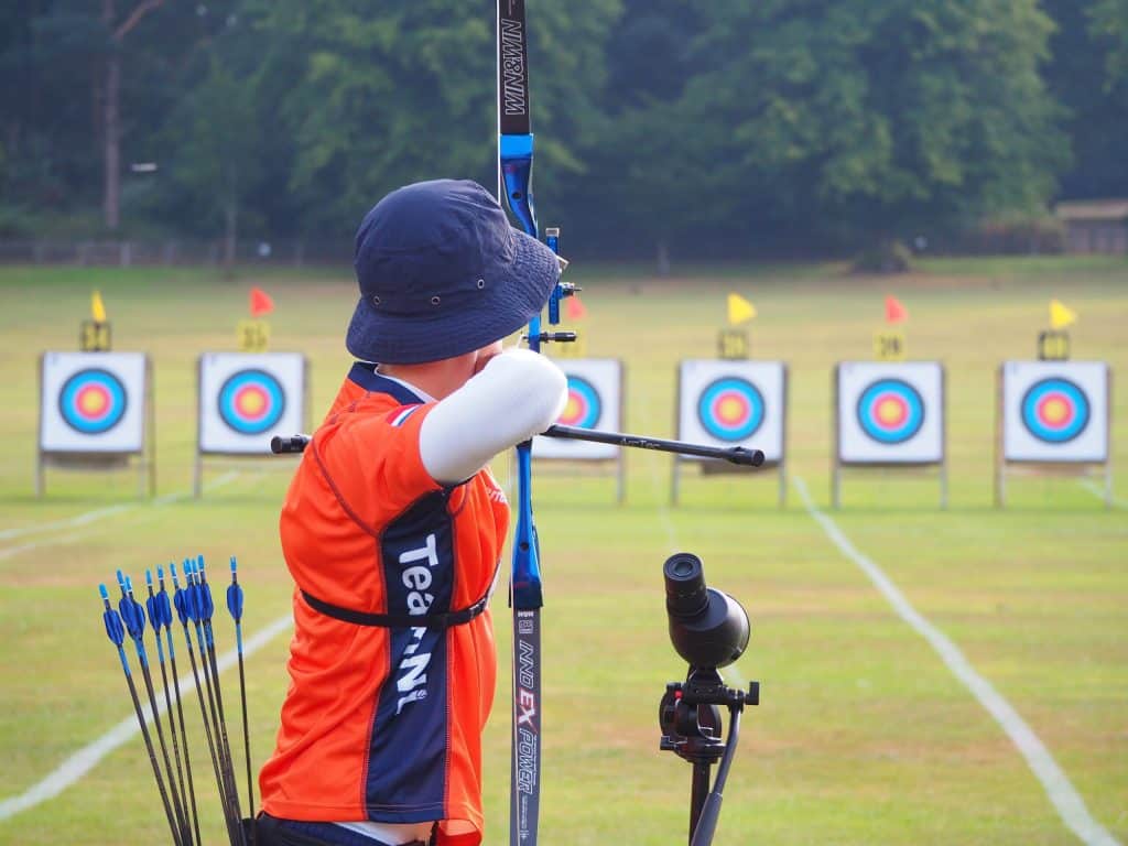 Een boogschutter in een oranje shirt richt zijn boog op een van de verschillende doelborden die op een veld zijn opgesteld. De doelen hebben veelkleurige roosjes en zijn genummerd. De boogschutter heeft een pijlenkoker op zijn rug, waarmee hij zijn passie voor handboogsport laat zien.