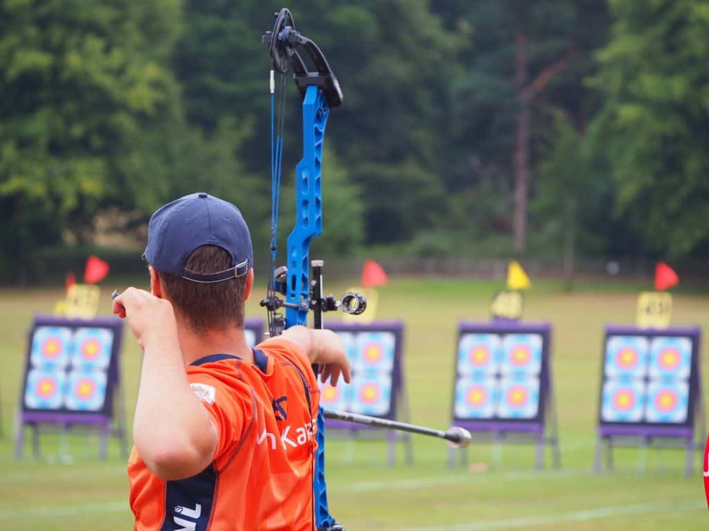 Een persoon die een oranje shirt en een pet draagt, richt een blauwe compoundboog op een rij boogschietdoelen die op een veld zijn opgesteld, waarmee hij zijn vaardigheden in de handboogsport laat zien.