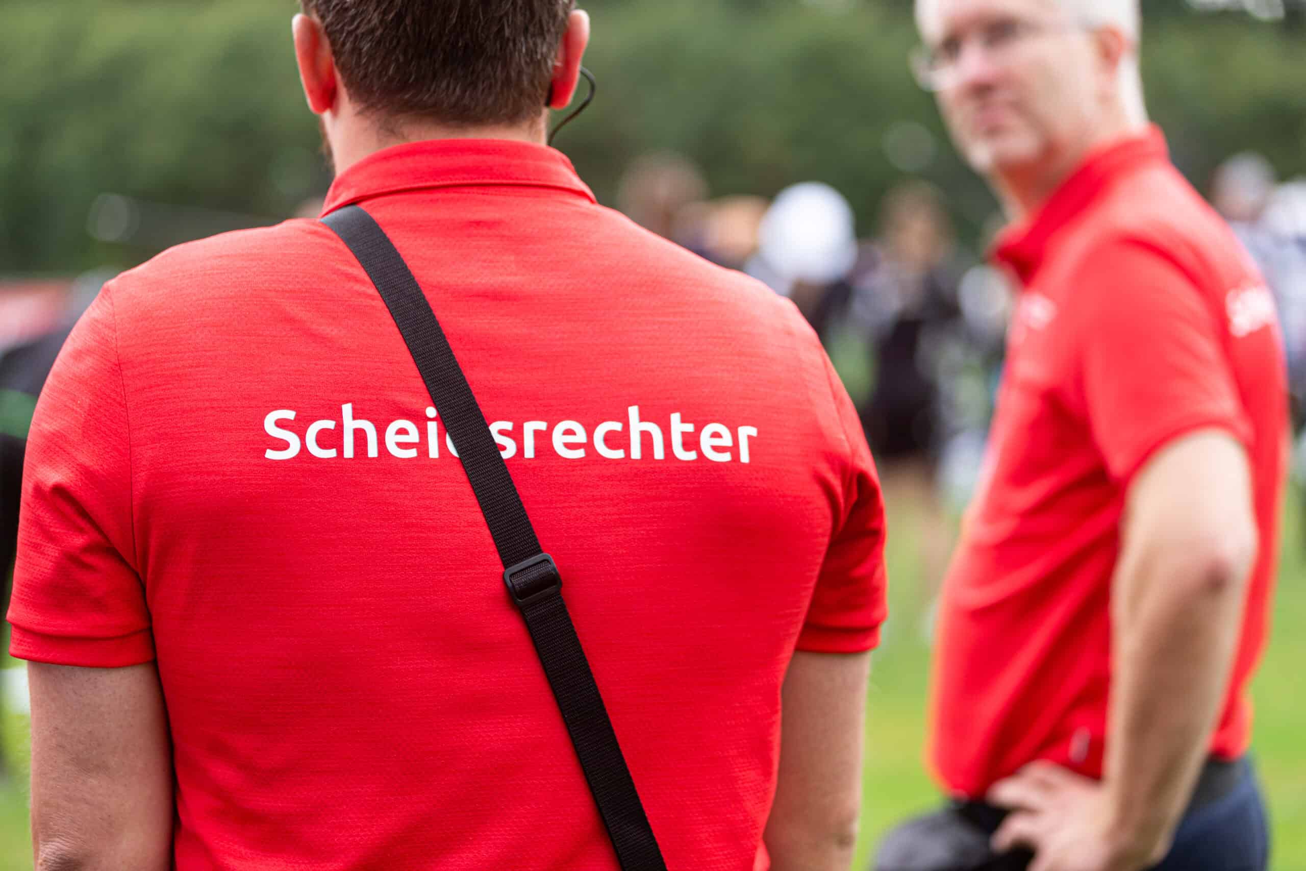 Twee personen die rode shirts dragen met de opdruk "Schiedsrichter" op de achterkant worden buiten gezien met een onscherpe achtergrond. De ene persoon kijkt de andere kant op, terwijl de ander zijwaarts draait, mogelijk als hij toezicht houdt op een handboogsportevenement.