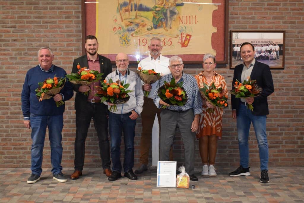 Zeven mensen staan voor een bakstenen muur met boeketten bloemen in hun handen. Een certificaat en een geschenkdoos worden bij hun voeten op de grond gelegd. Achter hen hangt, naast een groot ingelijst kunstwerk en een plaquette, een indrukwekkende pijl die wordt gebruikt voor handboogschieten.