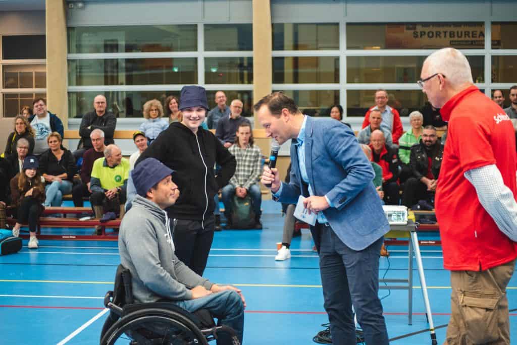 Een man in een rolstoel spreekt in een microfoon die wordt vastgehouden door een man in een blauwe blazer, met twee andere personen en een zittend publiek op de achtergrond, in een sporthal versierd met handboogsportapparatuur.