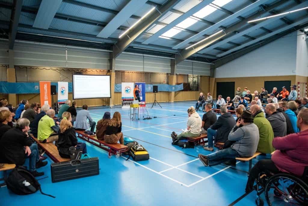 Een persoon presenteert informatie op een scherm aan een zittend publiek in een ruime kamer met een hoog plafond, vergelijkbaar met een boogschietbaan. De aanwezigen zitten op banken of stoelen, en sommige mensen staan.