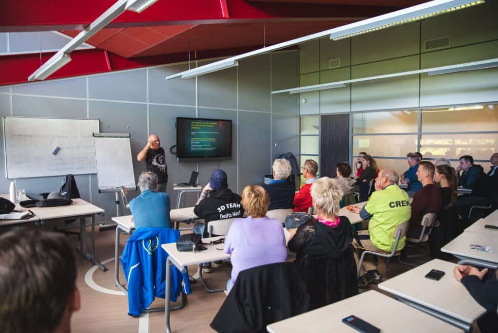 Een persoon staat vooraan in een klaslokaal en geeft een presentatie aan een groep zittende personen, die aandachtig lijken. Op een projectiescherm wordt kleurrijke tekst weergegeven die verband houdt met handboogsport, en verschillende mensen maken aantekeningen.