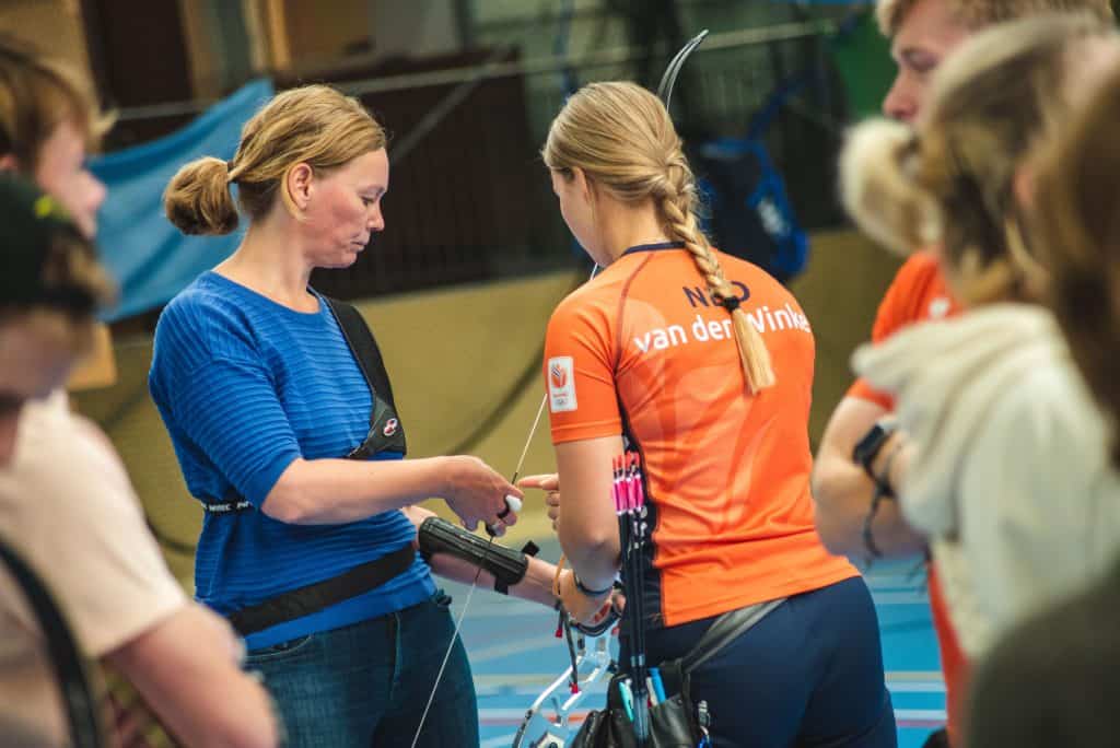 Een vrouw in een blauw shirt assisteert binnenshuis een boogschutter in een oranje shirt. Andere mensen staan in de buurt en observeren de handboogsportsessie. De boogschutter houdt een boog vast en heeft pijlen, ook wel pijl genoemd, in een pijlkoker die om haar middel is bevestigd.