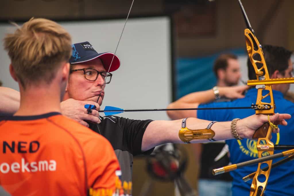 Een boogschutter met een pet en een bril richt zijn pijl en boog. Een andere persoon, met "NED" op zijn rug, staat op de voorgrond. Verschillende anderen staan op de achtergrond en vertegenwoordigen de handboogsportscene.