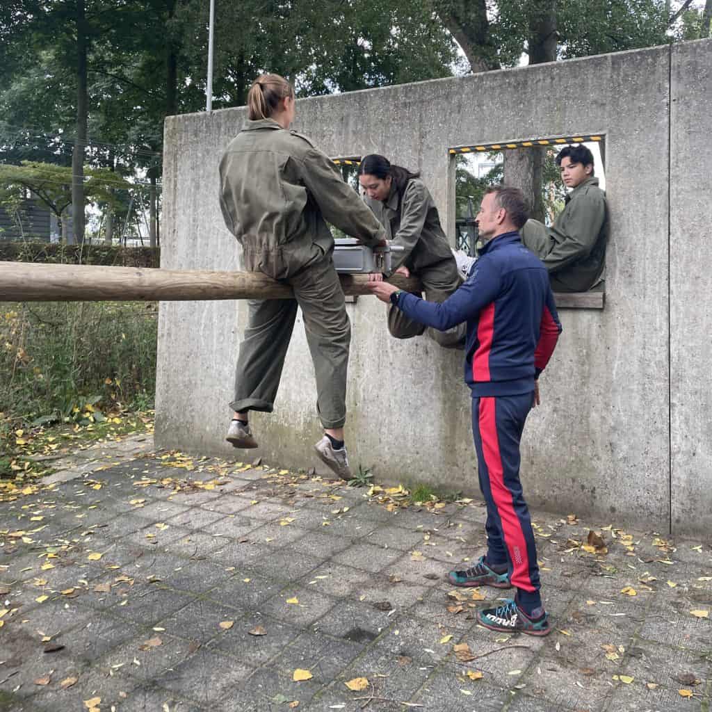 Vier personen, drie in olijfgroene outfits en één in een blauw en rood trainingspak, voeren buitenoefeningen uit op een betonnen constructie met houten balken en ramen in een parkomgeving, als onderdeel van hun handboogsporttraining.