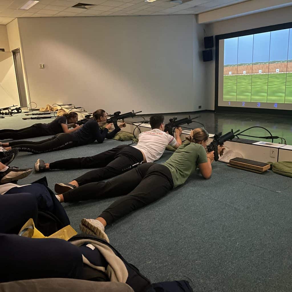 Een groep mensen ligt op de grond en richt hun geweren op een scherm waarop een schietbaandoel wordt weergegeven, net zoals boogschutters in de handboogsport mikken tijdens hun trainingssessies in een binnenfaciliteit.