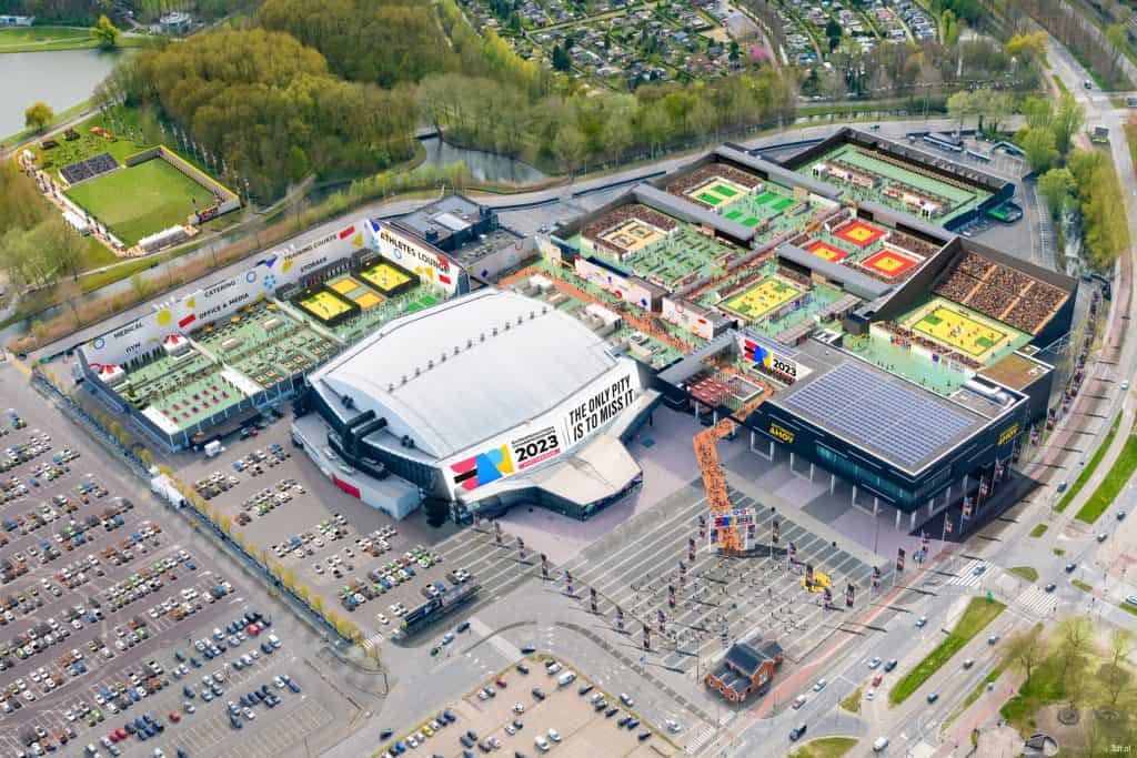 Luchtfoto van een groot multifunctioneel arenacomplex met buitensportvelden en parkeerplaatsen, met bewegwijzering voor de Pokémon Europe International Championships 2023, omgeven door groen en wegen. In de buurt is er een boogschietbaan voor liefhebbers van handboogsport.