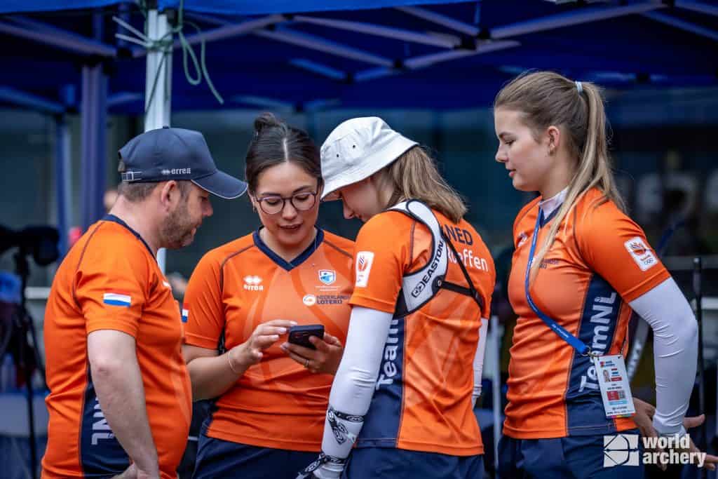 Vier boogschutters in oranje Team NL-uniformen staan samen onder een blauwe baldakijn, waarbij drie van hen gericht zijn op een smartphone die één van de boogschutters vasthoudt. Rechtsonder is een "World Archery" -logo zichtbaar, wat hun toewijding aan handboogsport benadrukt.