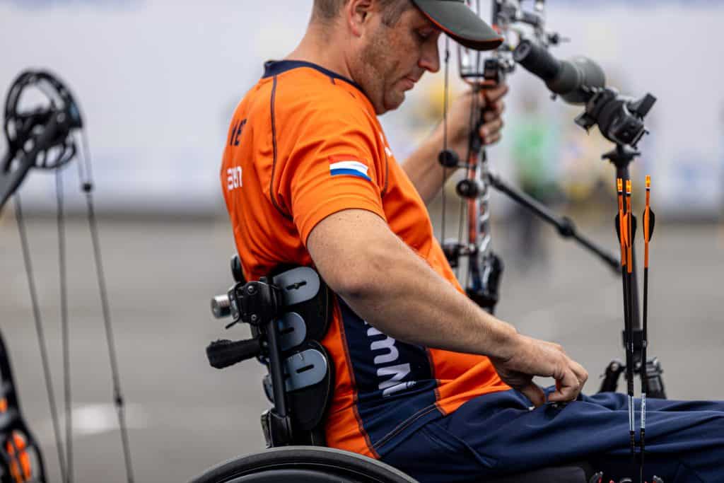 Een mannelijke boogschutter in een rolstoel, gekleed in een oranje shirt met een vlagpatch, bereidt zijn boog en pijlen voor op handboogsport tijdens een wedstrijd.