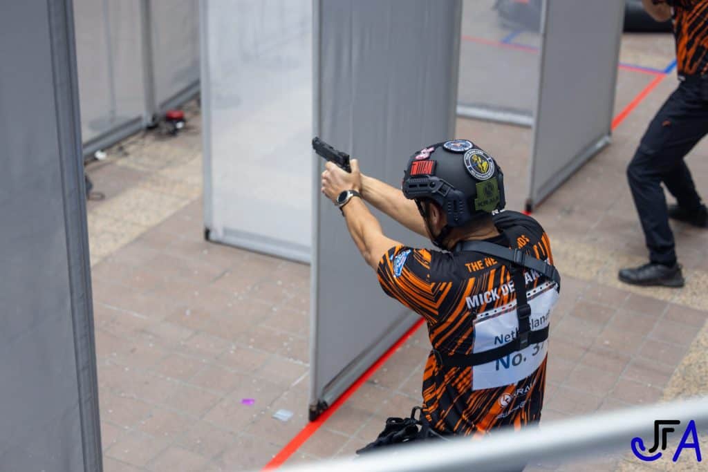 Een persoon met helm en oranje gestreept shirt richt een pistool op een overdekte schietbaan met scheidingswanden, wat doet denken aan de precisie van handboogschieten.