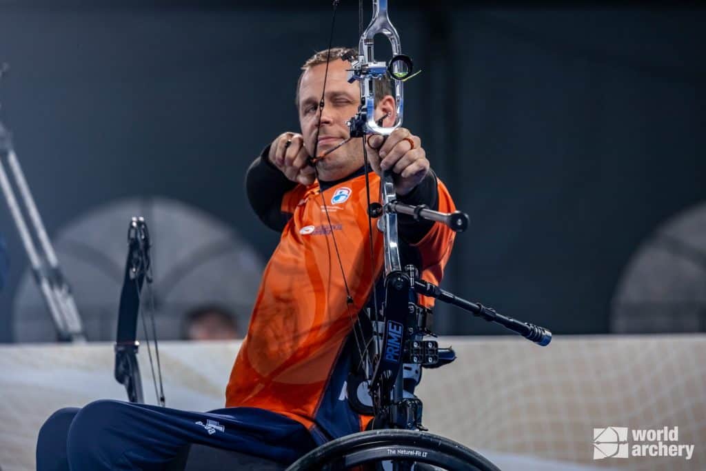 Een man in een oranje shirt richt een compoundboog terwijl hij in een rolstoel zit tijdens een handboogschietevenement. Rechtsonder in de afbeelding is het logo 'World Archery' zichtbaar.