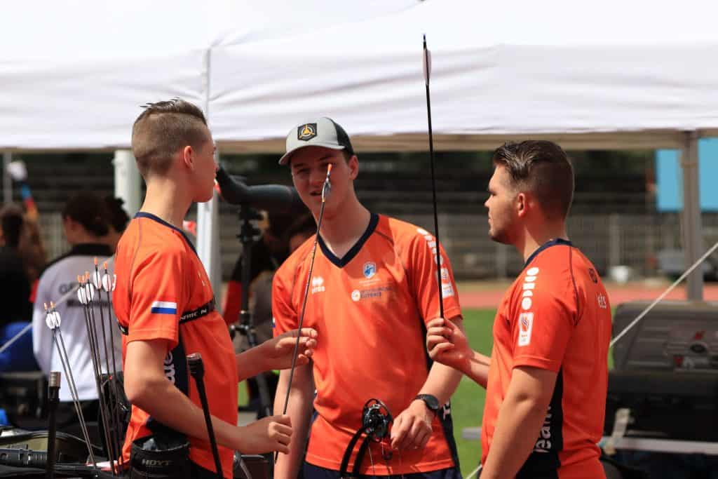 Drie mannelijke boogschutters in oranje shirts staan te praten terwijl ze hun pijl en boog vasthouden. Het lijkt erop dat ze een handboogschietevenement voorbereiden of bespreken.
