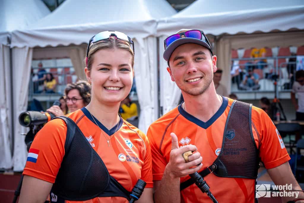 Twee boogschutters in bijpassende oranje uniformen staan naast elkaar glimlachend naar de camera. Rechtsonder in de afbeelding is een World Archery-logo te zien, dat hun passie voor handboogschieten laat zien.