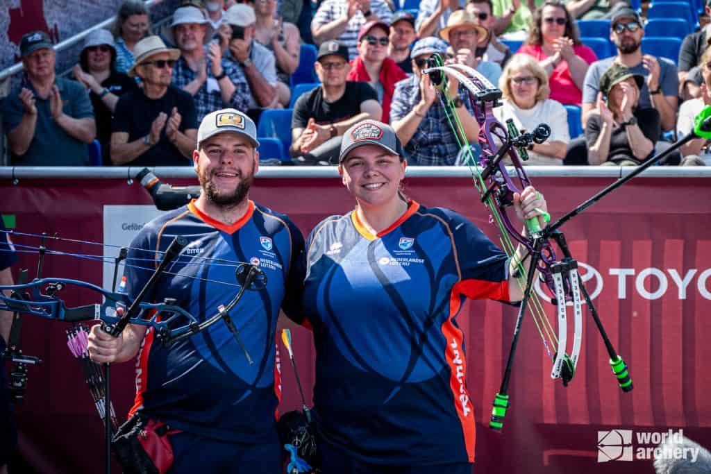 Twee boogschutters, een man en een vrouw, gekleed in bijpassende blauwe en rode uniformen, staan glimlachend met hun bogen in de handboogsport. Op de achtergrond klapt een publiek. Het logo "World Archery" is zichtbaar.