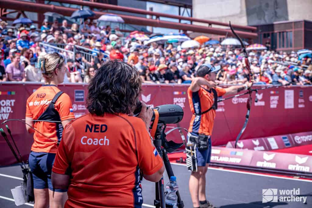 Boogschietcoach en teamleden uit Nederland observeren een deelnemer die op een doel mikt en schiet tijdens een handboogschietwedstrijd buiten met een grote menigte op de achtergrond.