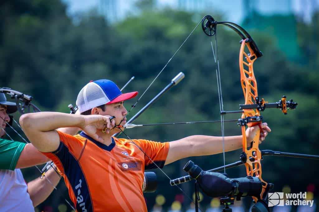 Een boogschutter in een oranje trui met een recurveboog mikt op een doelwit tijdens een handboogsportwedstrijd. Op de achtergrond zijn verschillende andere boogschutters zichtbaar. Het logo 'World Archery' staat rechtsonder.