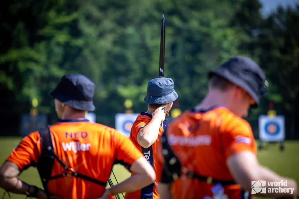 Drie boogschutters, gekleed in oranje shirts met Nederlandse namen en zwarte hoeden, bereiden zich voor om pijlen te schieten op verre doelen tijdens een handboogsportevenement in de open lucht.