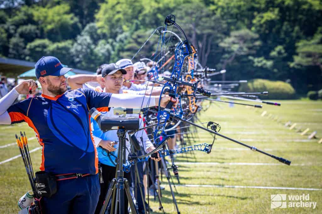 Boogschutters die in een rij staan, richten hun samengestelde bogen op doelen tijdens een handboogschietwedstrijd buiten. Op de voorgrond staat een mannelijke boogschutter in blauwe en oranje kleding. Logo van "World Archery" is zichtbaar.