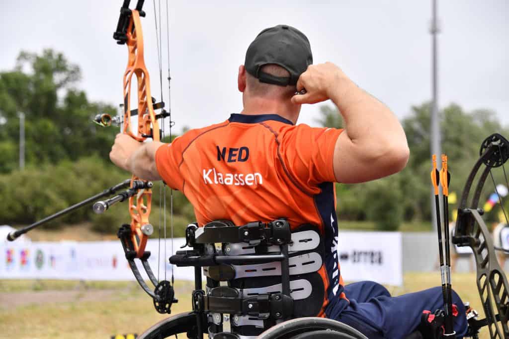 Een mannelijke boogschutter in een rolstoel bereidt zich voor om een pijl uit zijn boog te halen. Hij draagt een oranje shirt met op de rug "NED Klaassen", wat staat voor handboogschieten. Het evenement vindt buiten plaats met groen op de achtergrond.
