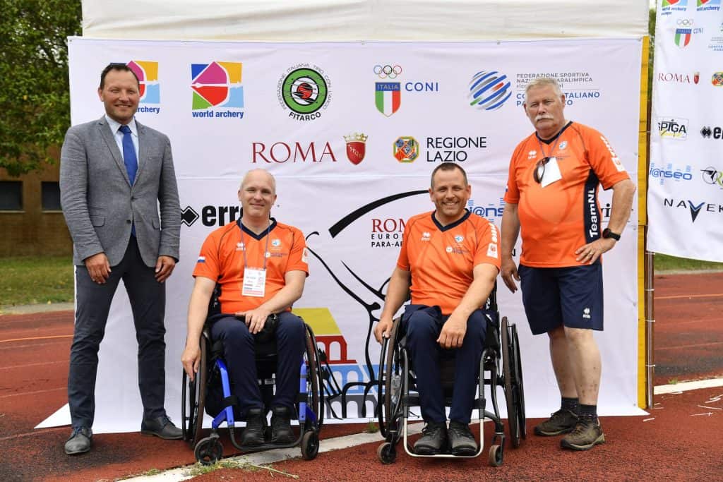 Vier mannen poseren voor een spandoek met verschillende logo's tijdens een sportevenement, waaronder twee zittend in een rolstoel in oranje sportkleding, en twee staande mannen - de een in pak en de ander in oranje sportkleding, waarmee ze hun toewijding aan handboogschieten laten zien.
