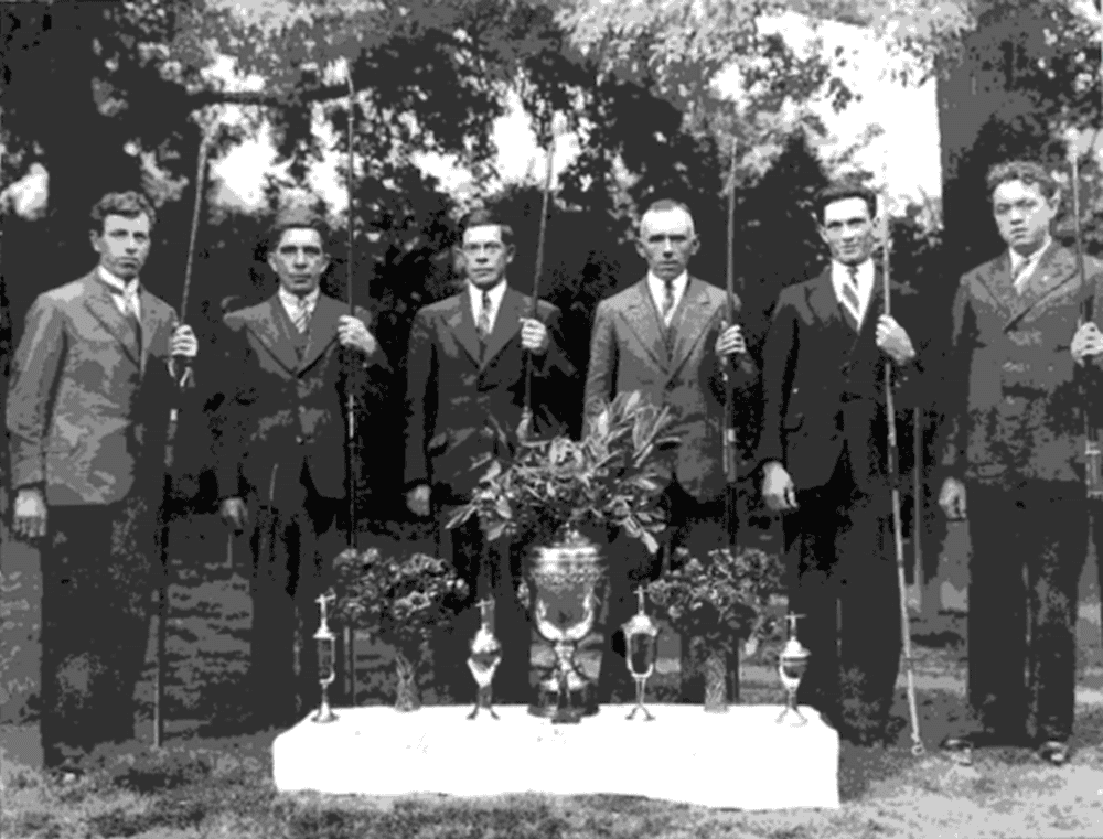 Een zwart-witfoto toont zes mannen in pak met golfclubs in hun handen, staande achter een tafel met trofeeën en bloemstukken, buiten met bomen op de achtergrond. De sfeer doet denken aan een formeel handboogsportevenement.
