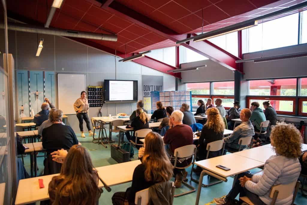 Een persoon geeft een presentatie aan een kamer met aandachtige personen die aan tafels zitten in een klaslokaal met natuurlijk licht dat door de ramen naar binnen valt, waarbij analogieën van handboogschieten worden gebruikt om de belangrijkste punten te illustreren.