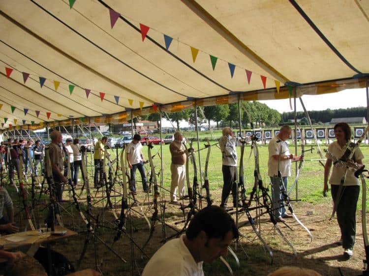 De deelnemers staan onder een tent met kleurrijke vlaggetjes en bereiden zich voor op het schieten met boogschieten op doelen aan de achterkant, waarbij ze de geest van handboogsport omarmen.