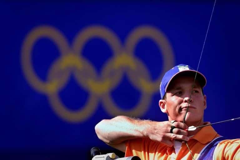 Een boogschutter in een oranje shirt en blauwe pet richt zijn boog, wat de precisie en vaardigheid van handboogschieten belichaamt, waarbij de Olympische ringen zichtbaar zijn op de vage blauwe achtergrond.