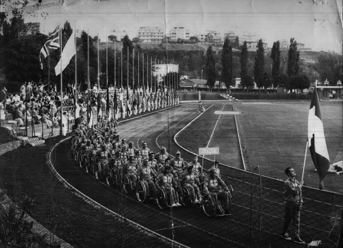 Een zwart-witfoto van een groep atleten in rolstoelen die deelnemen aan een parade op een atletiekbaan, mogelijk na een handboogschietevenement. Toeschouwers zitten op de tribunes en langs de baan hangen verschillende vlaggen.